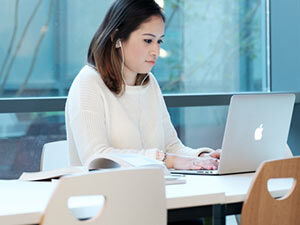 Malaysian female student sitting at a desk typing on a laptop with earphones in