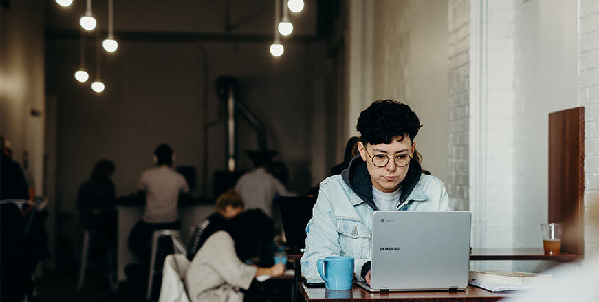 Malaysian male student sitting at a table, typing on a laptop with a cup beside him