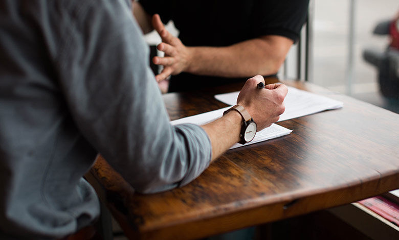 People sitting at a desk, paper on the desk, with their arms on the table one with a pen in his hand