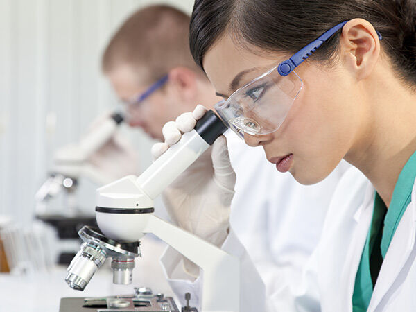 Female student wearing a lab coat and safety googles looking through a microscope